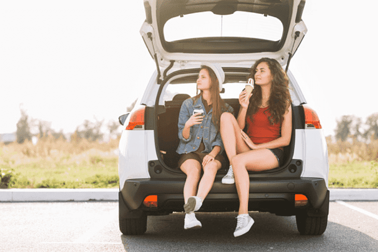 Jovens sentadas na bagageira do carro. Sugestões de refeições para viagens longas.